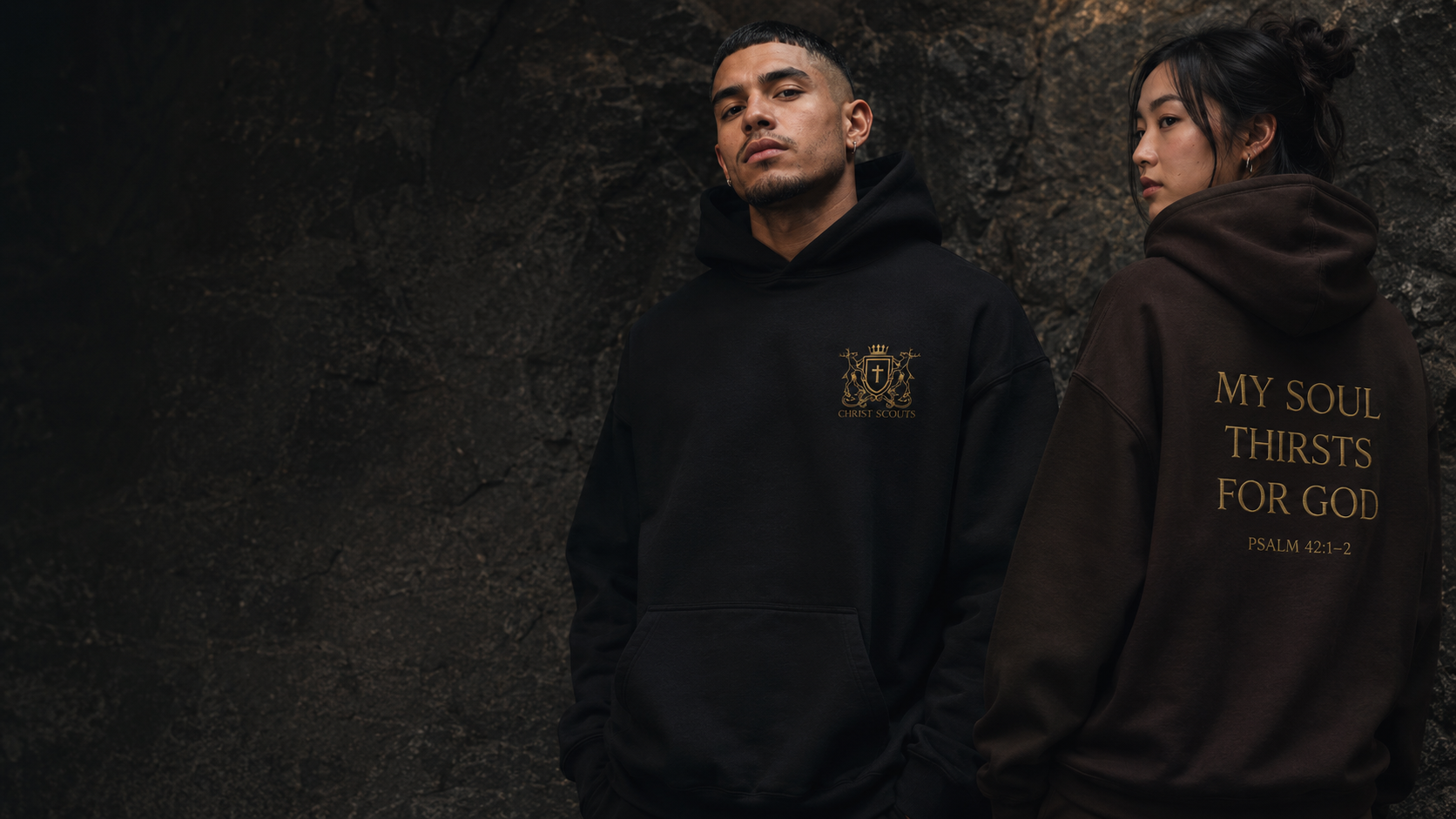 Two models wearing Christ Scouts hoodies against a dark textured wall — a man in a black hoodie and a woman in a dark brown hoodie in a cinematic luxury streetwear campaign image.